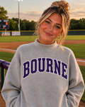 A woman wearing a light gray sweatshirt with 'BOURNE' printed in dark purple varsity letters, standing in front of a baseball field at sunset.