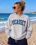 A woman wearing a light gray crewneck sweatshirt with 'POCASSET' printed in navy blue across the chest, standing on a sandy beach with the ocean and blue sky in the background.