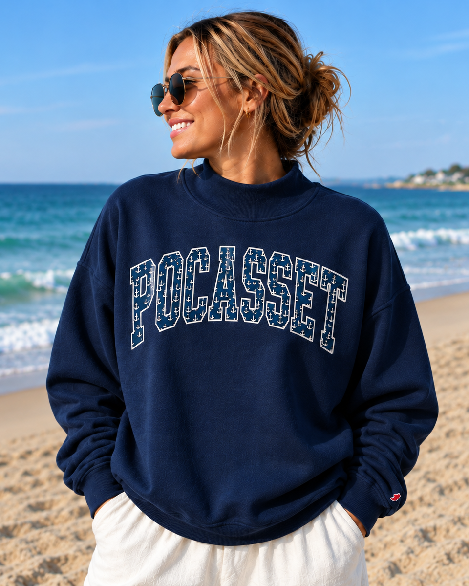 A woman wearing a navy blue crewneck sweatshirt with 'POCASSET' printed in white arched letters across the chest, standing on a sandy beach with the ocean and blue sky in the background.