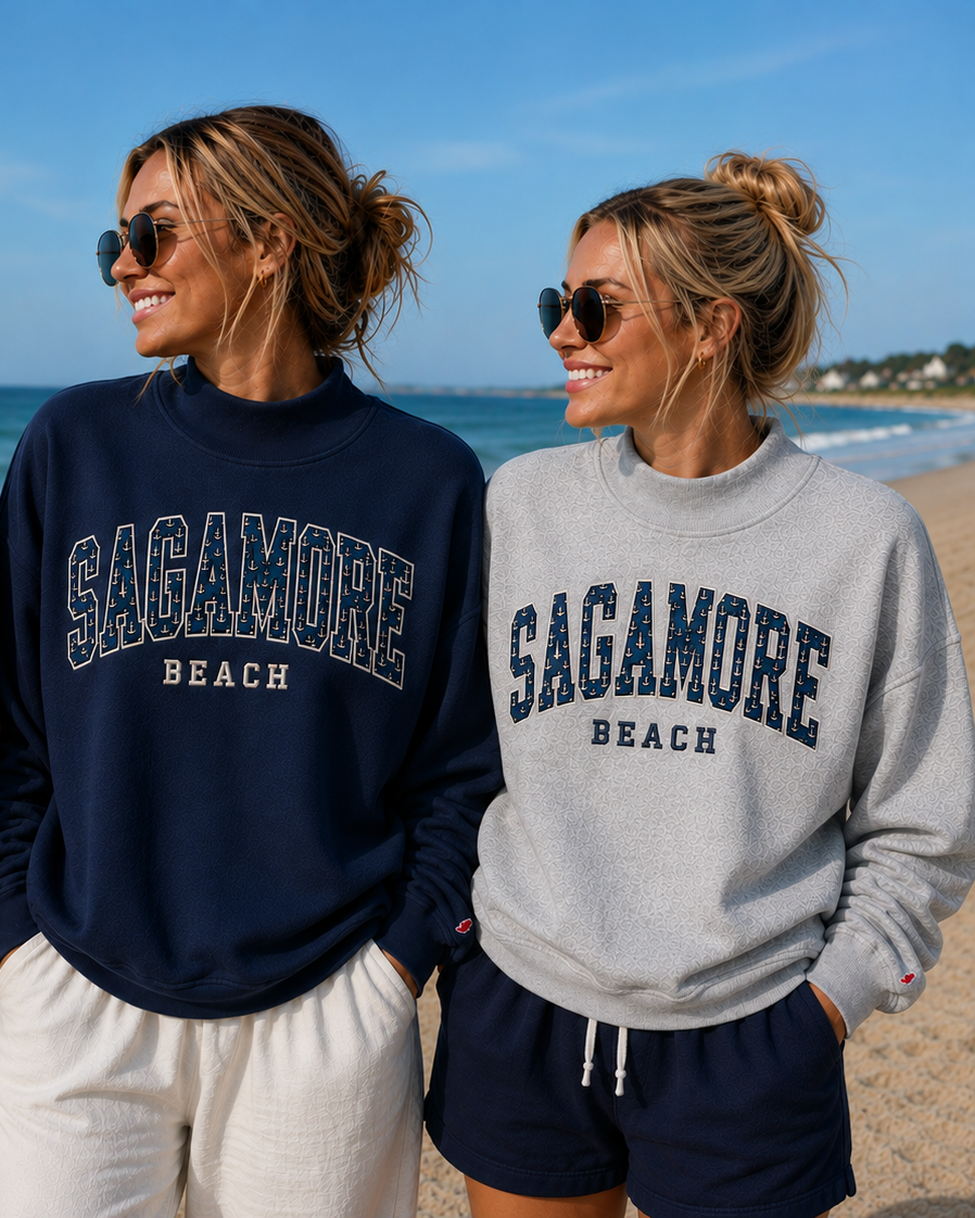 Two women standing on a beach wearing matching crewneck sweatshirts in red, navy, and heather gray, each with 'SAGAMORE BEACH' printed across the chest.